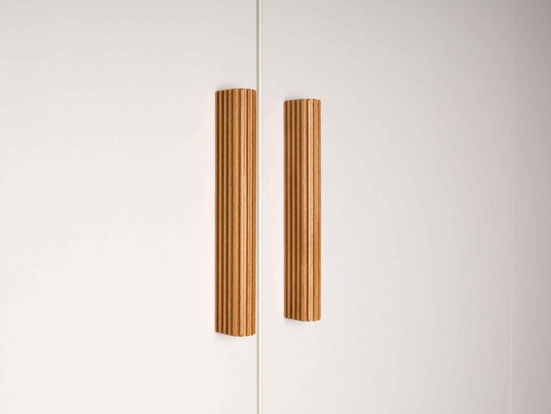 Op de afbeelding: Twee verticale, lichtbruine houten kastgrepen met een geribbeld ontwerp zijn gemonteerd op een witte kastdeur. De grepen zijn rechthoekig en hebben een warme, natuurlijke houttint. De kastdeur is gesloten.