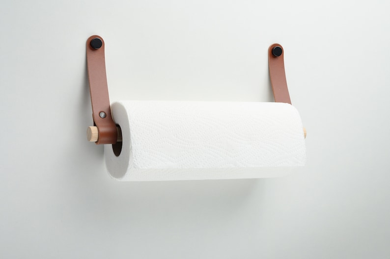 May include: A brown leather paper towel holder with wooden dowels and black metal hardware mounted on a white wall.