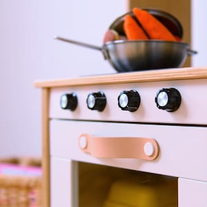 May include: A white toy stove with four black knobs and a tan leather handle. The stove has a glass oven door and a wooden top.