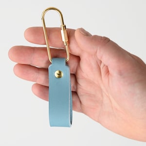Puede incluir: Un llavero de cuero azul claro con una correa de acabado liso y mate doblada en forma de bucle. La correa está asegurada con un remache de metal dorado y unida a un mosquetón de metal dorado grande y alargado con cierre de rosca.