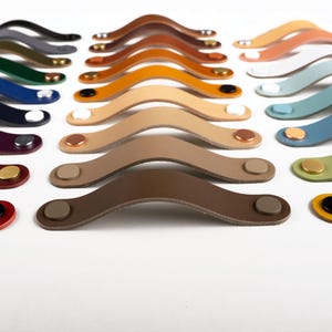 May include: A selection of leather cabinet pulls in various colours, including red, brown, beige, blue, green, and yellow. Each pull features a metal screw in the centre.