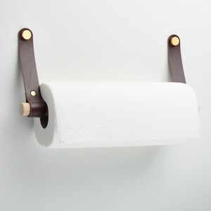May include: A brown leather paper towel holder with brass accents and a wooden dowel. The holder is mounted on a white wall.