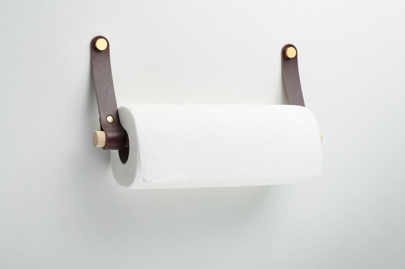 May include: A brown leather paper towel holder with brass accents and a wooden dowel. The holder is mounted on a white wall.