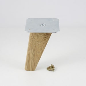 May include: A wooden furniture leg with a silver metal plate on top. The leg is angled and has a natural wood finish. The metal plate has four screw holes and a screw in the center.