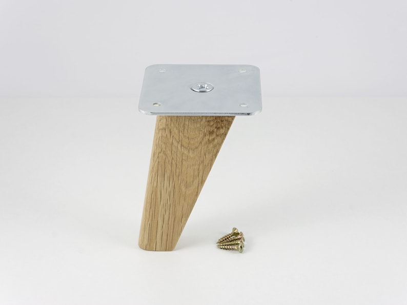 May include: A wooden furniture leg with a silver metal plate on top. The leg is angled and has a natural wood finish. The metal plate has four screw holes and a screw in the center.