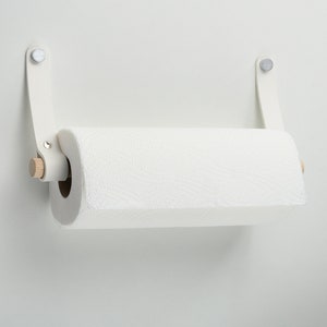 May include: White paper towel holder with a wooden dowel and white leather straps. The holder is mounted on a white wall.
