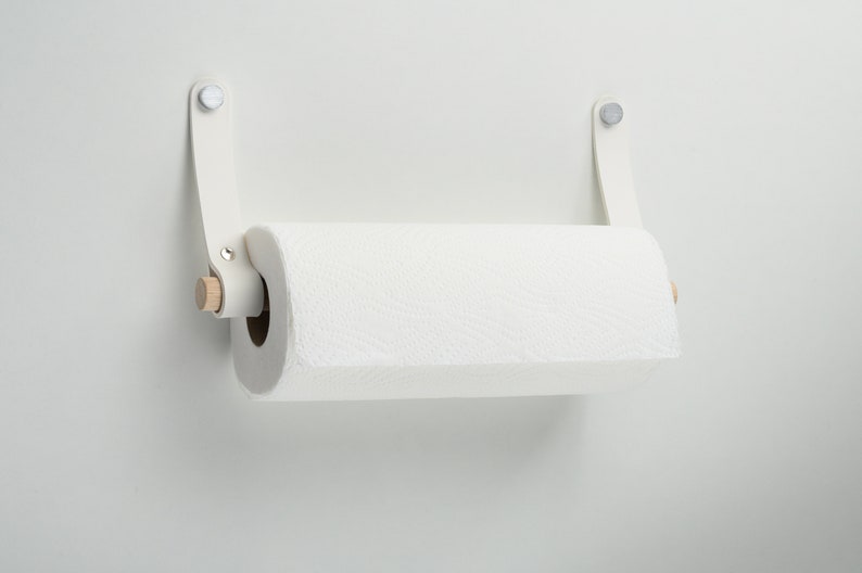 May include: White paper towel holder with a wooden dowel and white leather straps. The holder is mounted on a white wall.