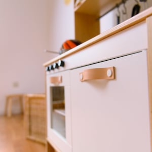 May include: White play kitchen with a brown leather handle and white knobs. The kitchen has a glass door oven and a white cabinet door.