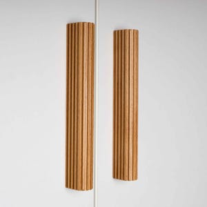 Op de afbeelding: Twee verticale houten kastgrepen met een geribbeld ontwerp. De grepen hebben een warme, natuurlijke houttint en zijn gemonteerd op een witte kastdeur. Het ontwerp is modern en minimalistisch.
