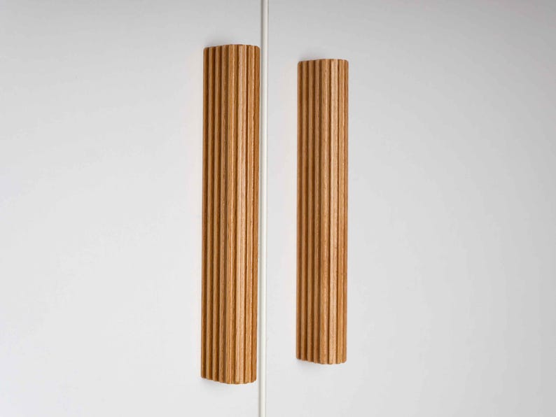 Op de afbeelding: Twee verticale houten kastgrepen met een geribbeld ontwerp. De grepen hebben een warme, natuurlijke houttint en zijn gemonteerd op een witte kastdeur. Het ontwerp is modern en minimalistisch.