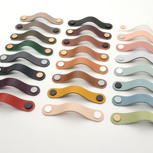 May include: Assortment of 18 leather cabinet pulls in various colors, including red, brown, green, blue, and gray. Each pull has a curved shape and two metal end caps.