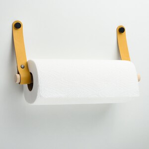 May include: A yellow leather paper towel holder with wooden dowels and black metal accents mounted on a white wall.