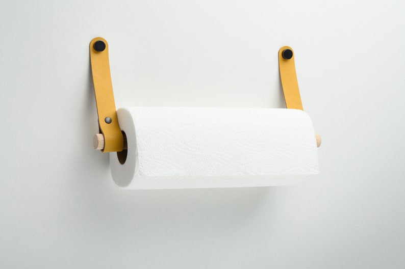 May include: A yellow leather paper towel holder with wooden dowels and black metal accents mounted on a white wall.