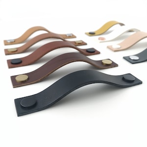 May include: Assortment of leather cabinet pulls in various colors, including brown, black, tan, and white. Each pull has a curved shape and features a metal screw.