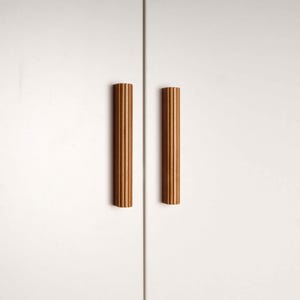 Op de afbeelding: Twee verticale, geribbelde houten kastgrepen in een warme bruine tint zijn gecentreerd tegen een witte achtergrond. De grepen zijn cilindrisch en hebben een reeks verticale groeven. De kastdeuren zijn gesloten.