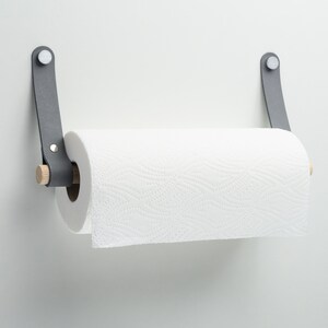 May include: A gray leather paper towel holder with a wooden dowel and a white paper towel roll. The holder is mounted on a white wall.