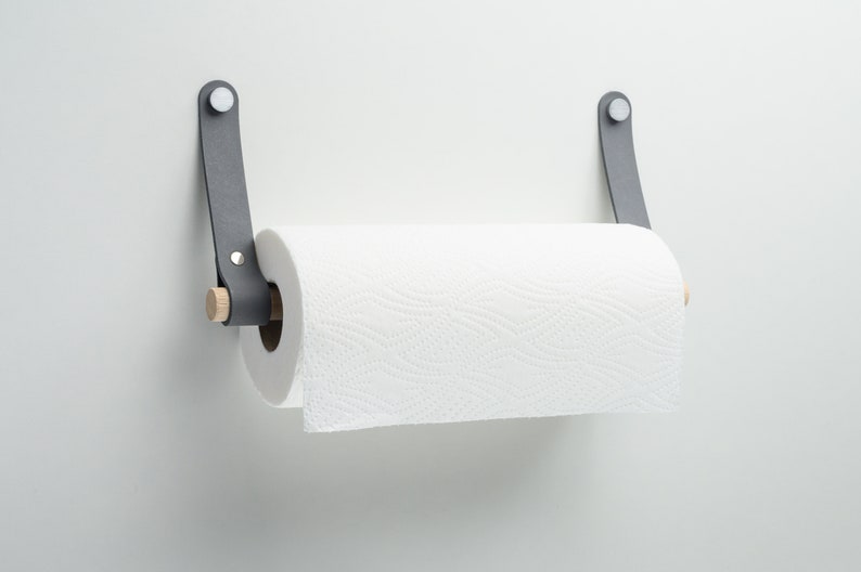 May include: A gray leather paper towel holder with a wooden dowel and a white paper towel roll. The holder is mounted on a white wall.
