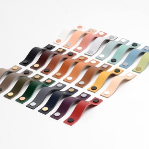 Op de afbeelding: Een selectie van leren kastgrepen in verschillende kleuren, gerangschikt in een verspringend patroon. De grepen hebben allemaal dezelfde vorm, met een gebogen ontwerp en twee metalen schroeven.