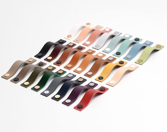 May include: A variety of leather cabinet pulls in different colors, arranged in a staggered pattern. The pulls are all the same shape, with a curved design and two metal screws.
