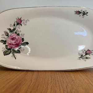 May include: An off-white rectangular serving platter with rounded corners and a gold-toned rim. The surface features a large pink rose floral arrangement on the left side and two smaller rose bud accents on the top right and bottom right.