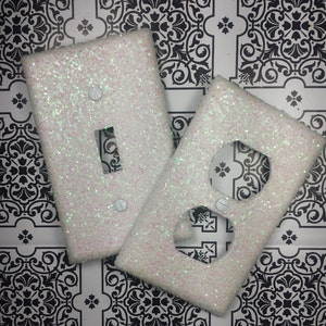May include: Two white glitter light switch and outlet covers. The covers are on a black and white patterned background.