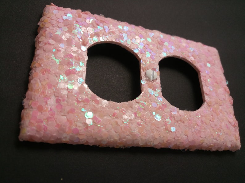 Puede incluir: Una cubierta decorativa de enchufe el&eacute;ctrico cubierta de purpurina rosa claro. La cubierta tiene dos aberturas para enchufes y un peque&ntilde;o tornillo blanco. La superficie est&aacute; cubierta de purpurina iridiscente.