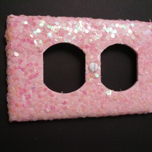 Puede incluir: Una cubierta de enchufe el&eacute;ctrica rosa, cubierta de purpurina, con dos aberturas circulares y un tornillo blanco. La cubierta es rectangular y tiene una superficie texturizada, con copos de purpurina iridiscentes.