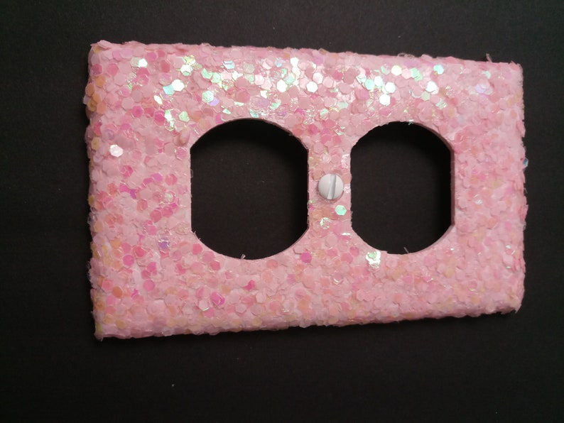 Puede incluir: Una cubierta de enchufe el&eacute;ctrica rosa, cubierta de purpurina, con dos aberturas circulares y un tornillo blanco. La cubierta es rectangular y tiene una superficie texturizada, con copos de purpurina iridiscentes.