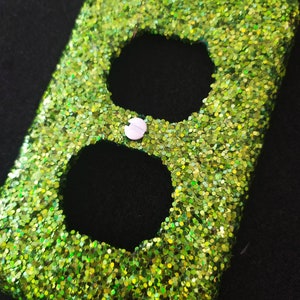 May include: A green glitter outlet cover with two round openings. The cover has a white screw in the center.