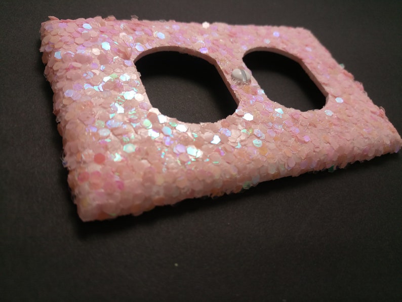 Puede incluir: Una placa de interruptor de luz rosa cubierta de purpurina con dos aberturas. La placa es rectangular con bordes redondeados y est&aacute; densamente cubierta de purpurina hexagonal iridiscente. Se ve un peque&ntilde;o elemento decorativo blanco.