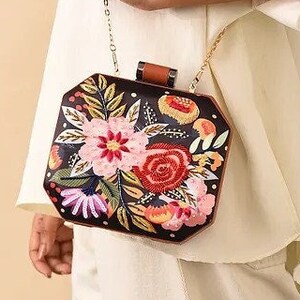 May include: A black and brown floral embroidered clutch bag with a gold chain strap. The bag has a geometric shape and features a variety of colorful flowers.