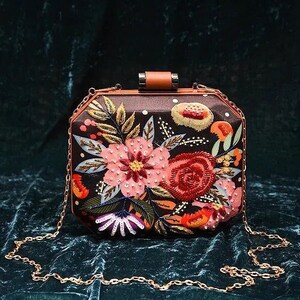 May include: A black and brown floral embroidered clutch bag with a gold chain strap. The bag has a geometric shape and features a variety of colorful flowers and leaves.