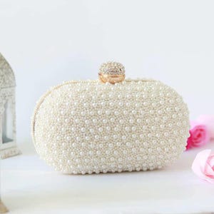 May include: Ivory-colored clutch covered in small, round faux pearls. The oval-shaped bag has a gold-colored clasp and a decorative, jeweled closure. The background is white with a hint of pink flowers.