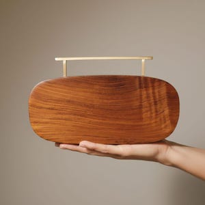 Puede incluir: Un bolso de mano de madera hecho a mano con forma rectangular y bordes redondeados. El bolso presenta un patrón de vetas de madera natural y un asa dorada. El bolso se sostiene en la mano de una persona sobre un fondo neutro.