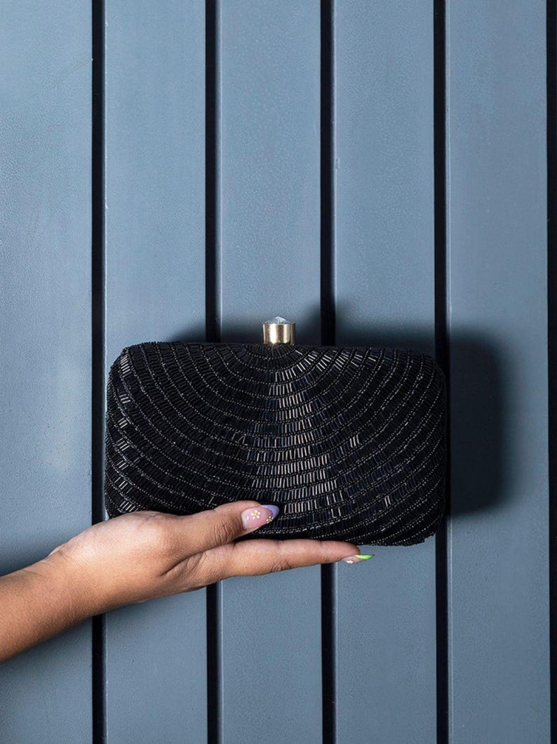 Puede incluir: Un clutch negro de cuentas con un cierre dorado. El clutch es peque&ntilde;o y rectangular.