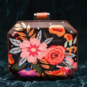 May include: A black clutch purse with a floral print featuring pink, red, orange, and purple flowers on a dark background. The purse has a brown top handle and a silver clasp.