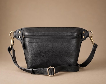 May include: A black pebbled leather belt bag featuring a horizontal zippered main compartment, antique brass-toned metal hardware, and an adjustable strap with a sliding buckle. The bag is secured with side rivets and metal D-ring attachments.