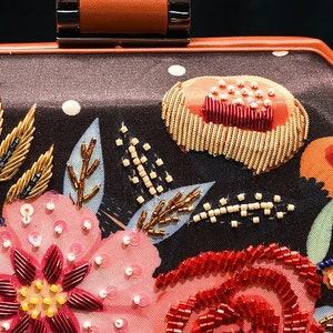 May include: A black satin clutch bag with a floral design. The flowers are embroidered with sequins and beads in shades of pink, red, and gold. The bag has a brown leather top handle.