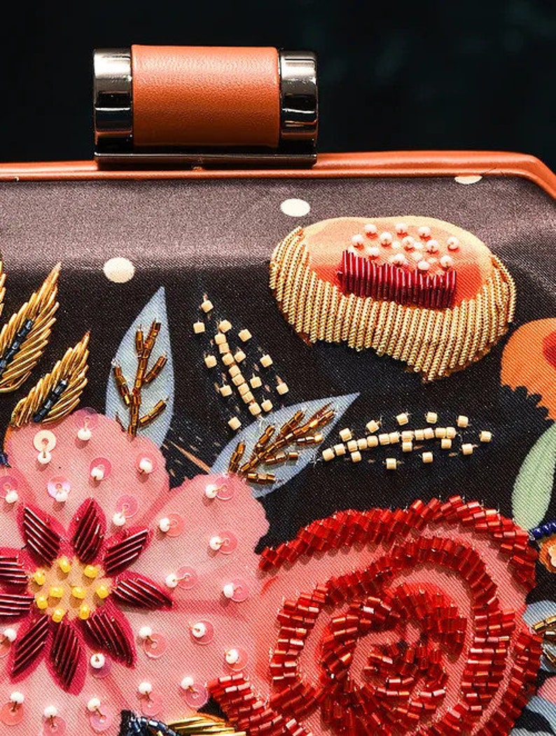 May include: A black satin clutch bag with a floral design. The flowers are embroidered with sequins and beads in shades of pink, red, and gold. The bag has a brown leather top handle.