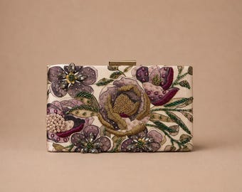 Può includere: Una pochette rettangolare con base bianco sporco e intricati ricami floreali nelle tonalità del viola, oro e verde. Il design presenta dettagli in perline, fili metallici e una chiusura a scatto in metallo dorato sulla parte superiore.