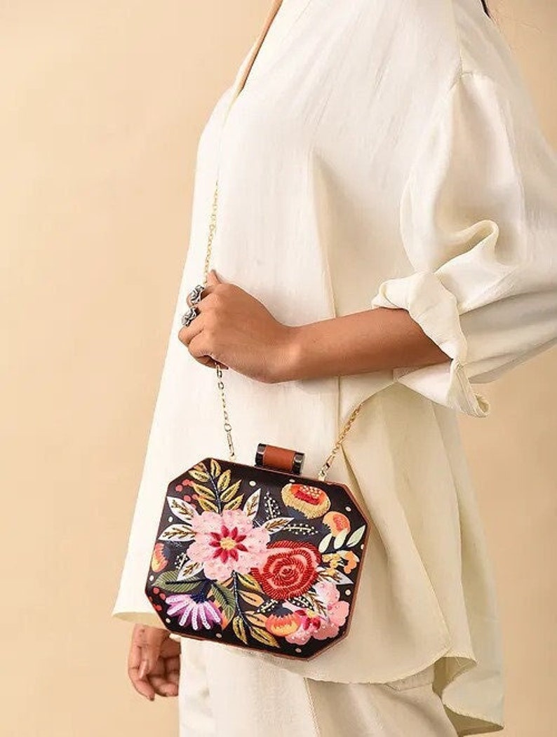 May include: A black and brown floral embroidered clutch bag with a gold chain strap. The bag has a geometric shape and features a variety of colorful flowers.