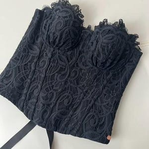 Puede incluir: Un top tipo corsé negro sin tirantes con un intrincado encaje floral superpuesto, copas moldeadas estructuradas con ribete de encaje festoneado en el escote, detalles de varillas verticales y largas cintas negras en el dobladillo.