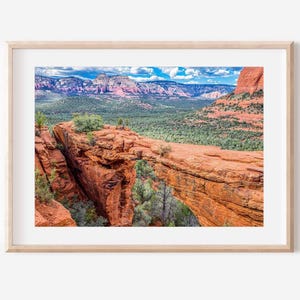 May include: A framed landscape photograph featuring a natural red rock sandstone bridge formation in a desert canyon. The scene includes green vegetation, distant layered mountains, and a bright blue sky with white clouds, all encased in a light wood frame.