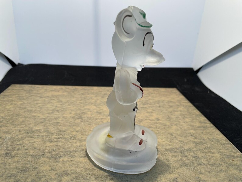 Puede incluir: Una figurita de vidrio transparente de un personaje de dibujos animados con un sombrero. La figurita est&aacute; de pie sobre una base de vidrio transparente.