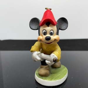 Puede incluir: Una figurita de cer&aacute;mica de Mickey Mouse con un sombrero rojo y una chaqueta amarilla, sosteniendo un rollo en sus manos. La figurita est&aacute; de pie sobre una base verde.