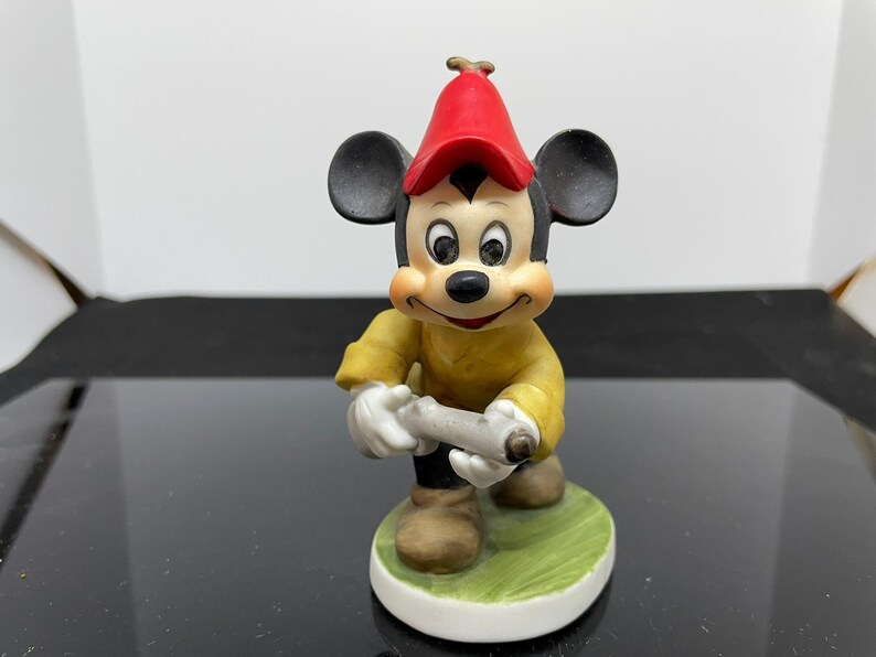 Puede incluir: Una figurita de cer&aacute;mica de Mickey Mouse con un sombrero rojo y una chaqueta amarilla, sosteniendo un rollo en sus manos. La figurita est&aacute; de pie sobre una base verde.