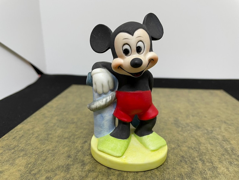 May include: A ceramic Mickey Mouse figurine. Mickey is wearing red shorts, black shoes, and green shoes. He is leaning against a blue and gray post. The figurine is on a yellow base.