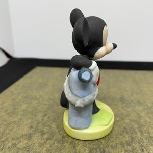 May include: A ceramic figurine of Mickey Mouse, wearing a blue and white outfit with a red heart. The figure is standing on a yellow base with green grass. The figurine has black ears and a black head.