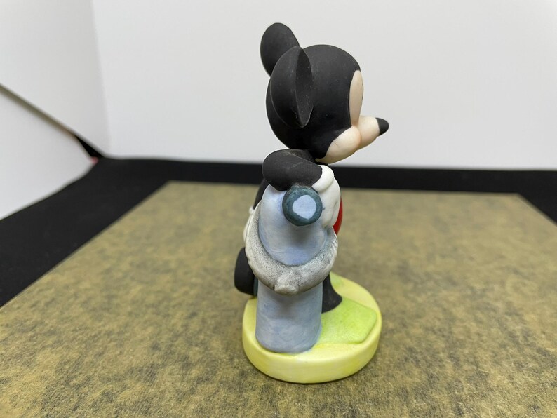 May include: A ceramic figurine of Mickey Mouse, wearing a blue and white outfit with a red heart. The figure is standing on a yellow base with green grass. The figurine has black ears and a black head.