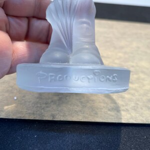 May include: A clear glass figurine of a person wearing a hat and boots. The figurine is standing on a base that says "Productions".